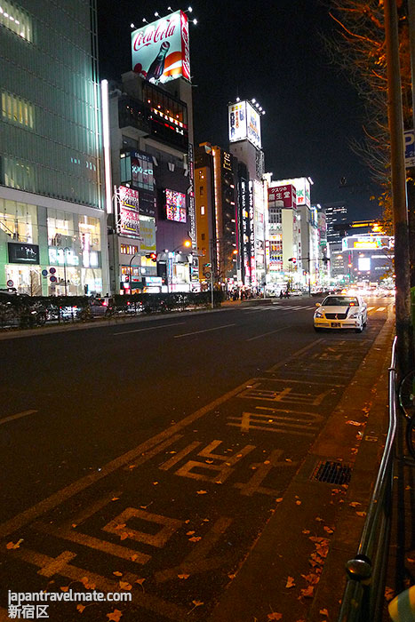 A street leading up to Shinjuku Station, lots of cars and shops and electronic signs.